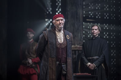Shylock in bluish and redish robes, burgundy vest and white shirt plus red skullcap and hands behind his back, Prince in black robe behind him, guard in leather waiste coat and red breeches, light streaming through the grate of the back wall.